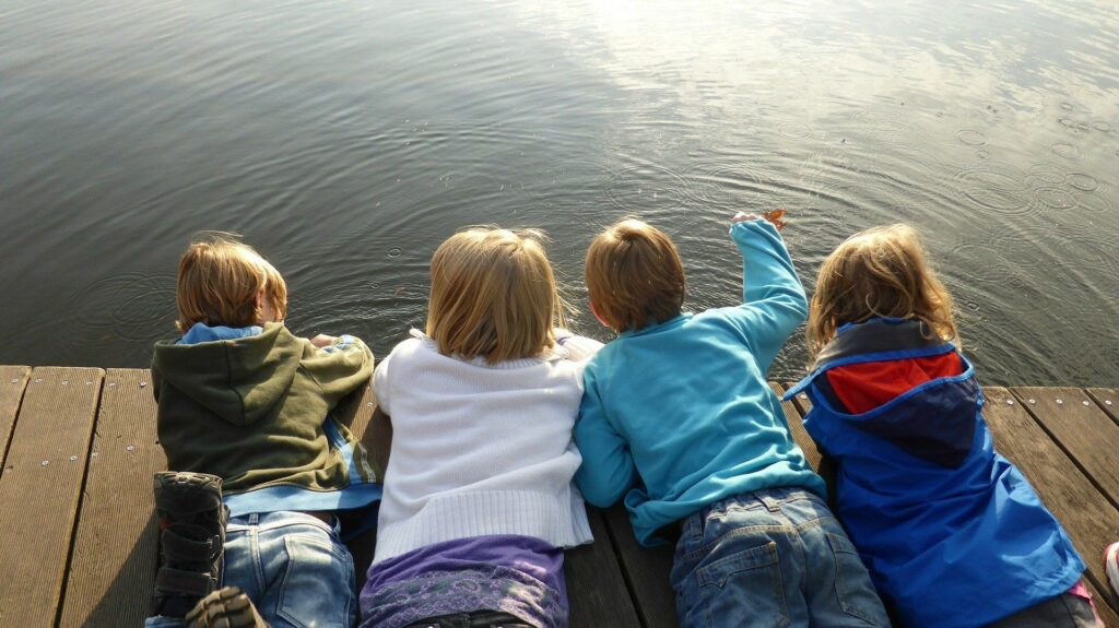 Four children laying on a wooden bridge looking at a lake reflecting the warm sunlight